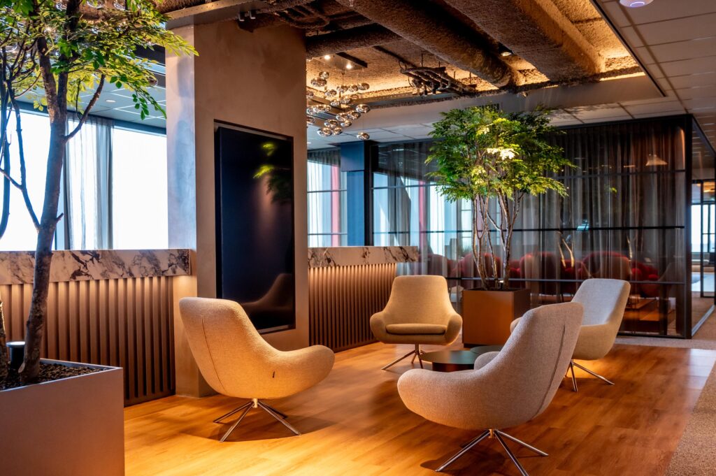 Moderne loungehoek met vier beige designstoelen, houten vloer, grote kamerplanten en sfeervolle verlichting in een kantoor aan de P.J. Oudweg.